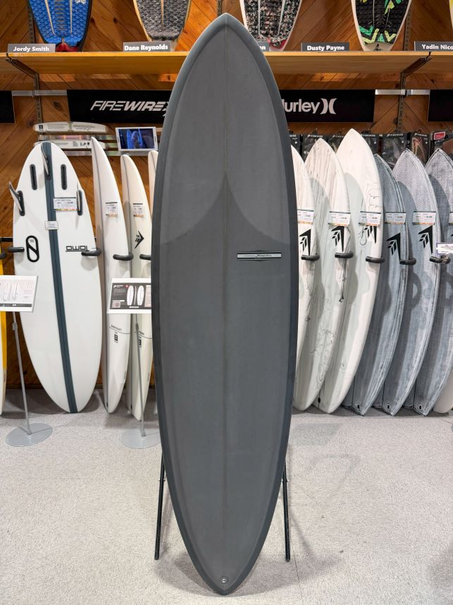 IMG_8980 - SHOP NEWS | ムラサキスポーツオンラインストア 通販 Large gray surfboard on a stand in a surf shop, with white and patterned boards lining the wall behind.