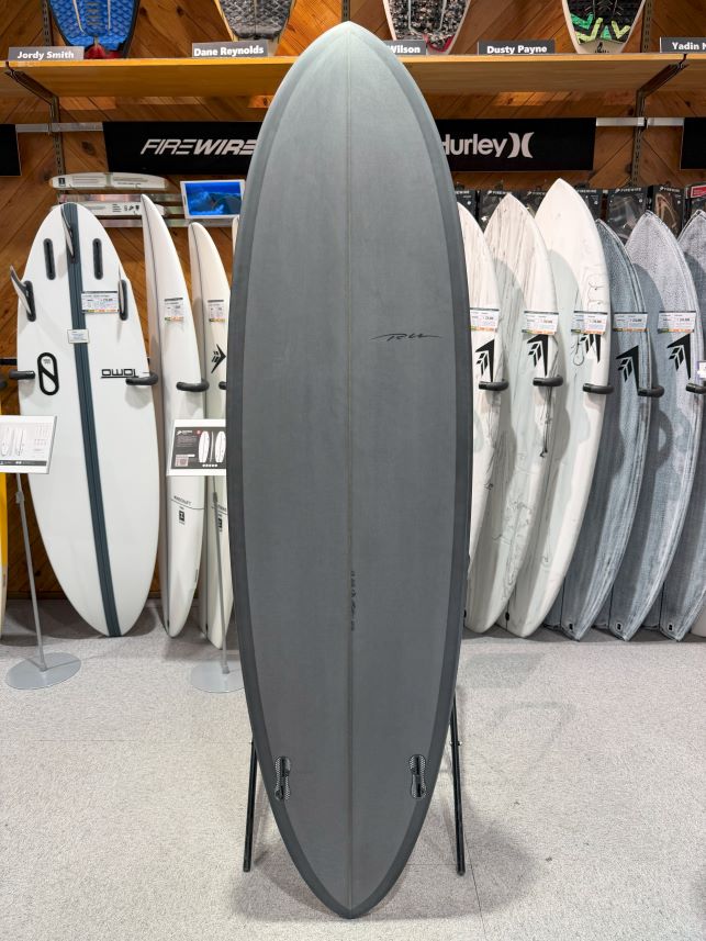 IMG_8981 - SHOP NEWS | ムラサキスポーツオンラインストア 通販 Gray surfboard on a stand in a surf shop, with a row of boards on shelves behind it.