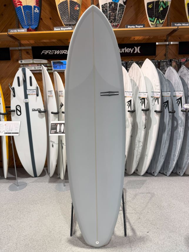 IMG_8984 - SHOP NEWS | ムラサキスポーツオンラインストア 通販 Tall light-gray surfboard on a stand in a shop, surrounded by other boards and racks in the background.