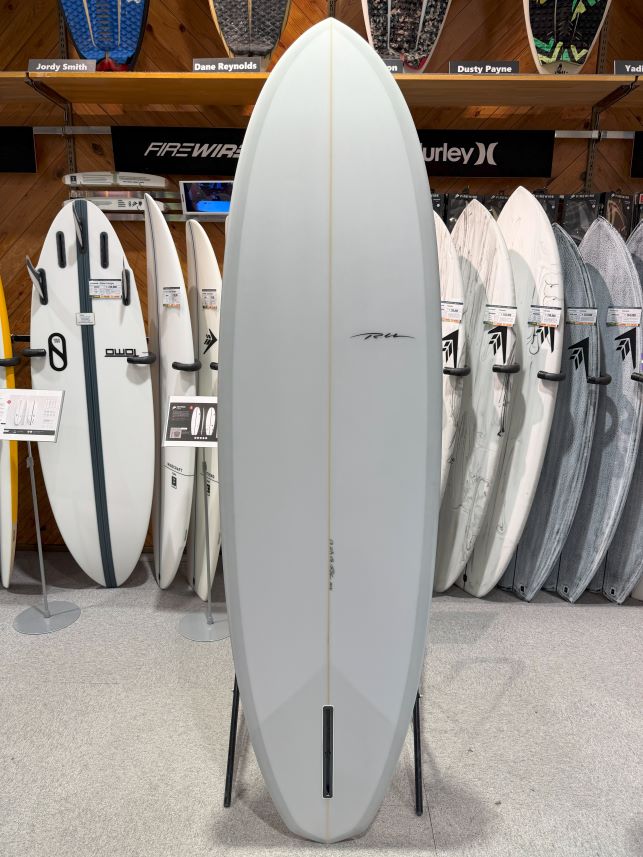 IMG_8985 - SHOP NEWS | ムラサキスポーツオンラインストア 通販 Tall white surfboard standing upright on a stand in a shop, surrounded by other boards on racks.