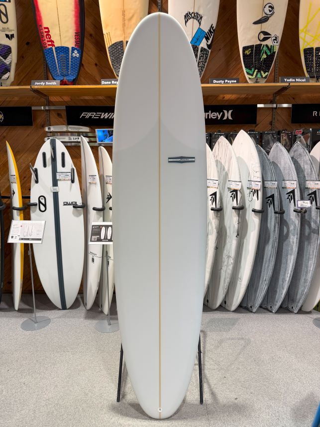 IMG_8989 - SHOP NEWS | ムラサキスポーツオンラインストア 通販 Tall white surfboard on display stand in a shop, surrounded by other boards on racks and wall shelves.
