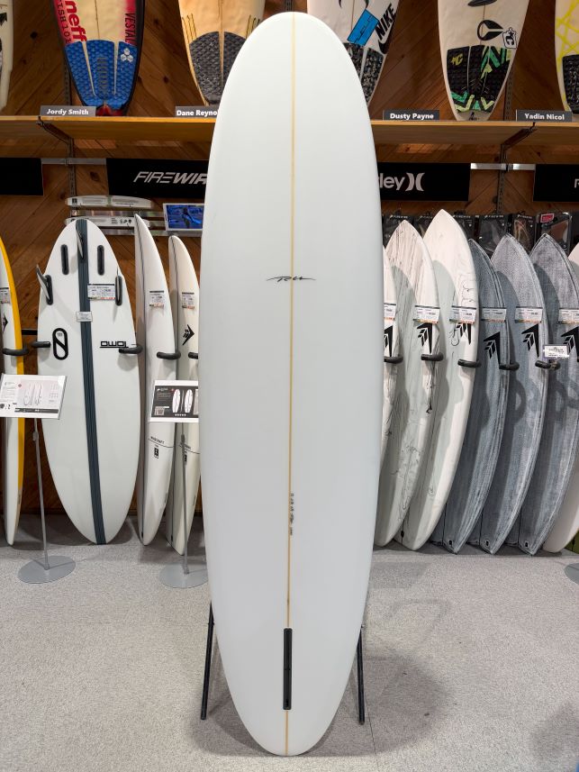 IMG_8990 - SHOP NEWS | ムラサキスポーツオンラインストア 通販 White surfboard displayed upright on a stand in a store, featuring a single beige stringer down the center and surrounded by other boards on racks.