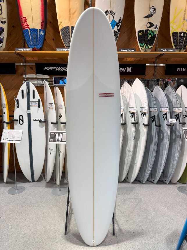 IMG_8992 - SHOP NEWS | ムラサキスポーツオンラインストア 通販 White surfing board on a stand in a shop, surrounded by other boards on racks and on the wall displays behind it.
