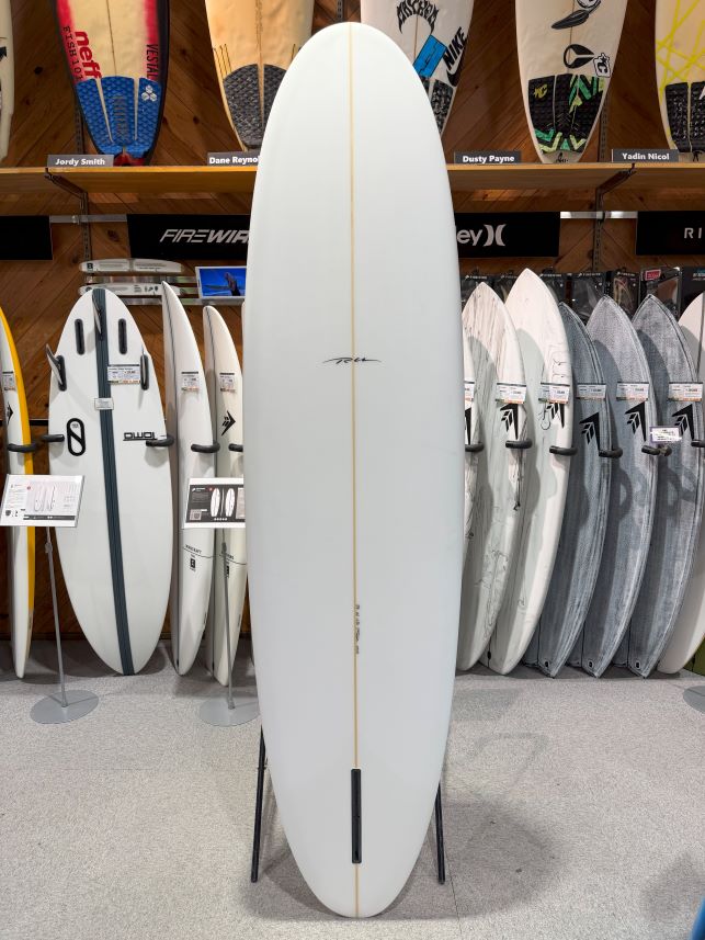 IMG_8993 - SHOP NEWS | ムラサキスポーツオンラインストア 通販 White surfboard on a stand in a shop, centered among other boards, with a narrow wood stringer running lengthwise and a small fin box near the tail.