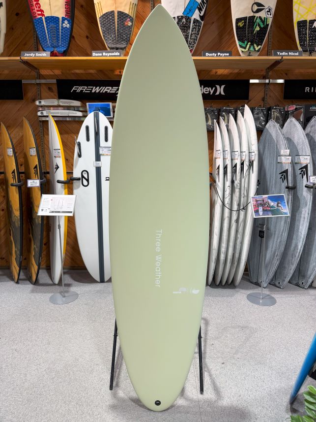 IMG_9403 - SHOP NEWS | ムラサキスポーツオンラインストア 通販 Pale green surfboard standing upright on a black stand in a surf shop, with racks of other boards and paddles visible on the walls behind it.
