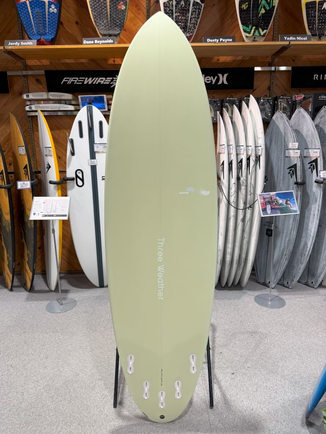 IMG_9404 - SHOP NEWS | ムラサキスポーツオンラインストア 通販 Pale green surfboard on a stand inside a surf shop, with racks of boards in the background.