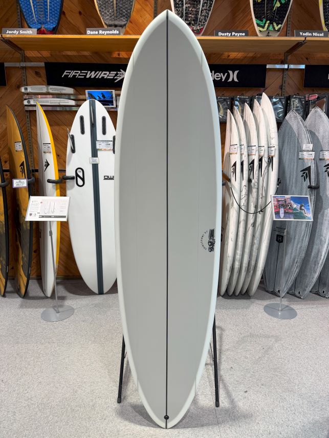 IMG_9432 - SHOP NEWS | ムラサキスポーツオンラインストア 通販 Tall gray surfboard on a display stand in a surf shop, with other boards on racks behind it.