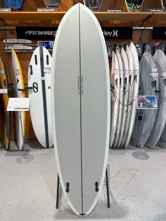 IMG_9433 - SHOP NEWS | ムラサキスポーツオンラインストア 通販 Light gray surfboard on a stand in a surf shop, with other boards displayed on racks in the background.