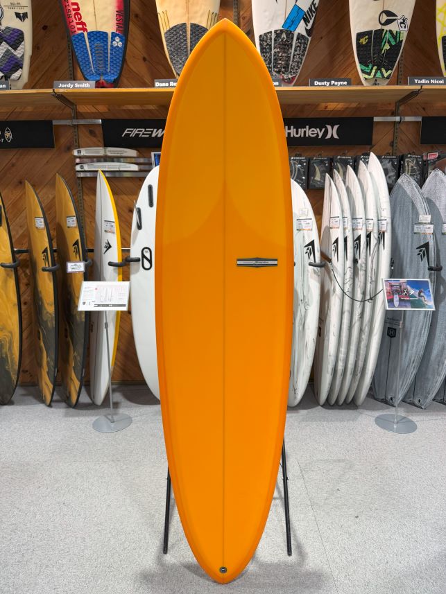 IMG_9508 - SHOP NEWS | ムラサキスポーツオンラインストア 通販 Orange surfboard standing upright on a display stand in a surf shop, with many other boards on racks in the background.