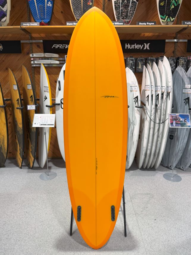 IMG_9509 - SHOP NEWS | ムラサキスポーツオンラインストア 通販 Bright orange shortboard standing upright on a display stand in a surf shop, with other boards visible in the background.