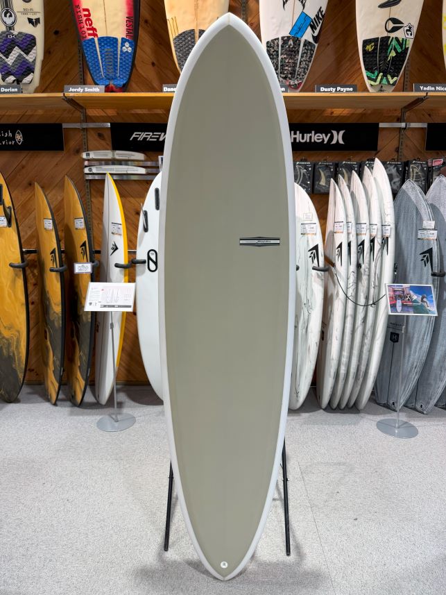 IMG_9512 - SHOP NEWS | ムラサキスポーツオンラインストア 通販 Tall gray surfboard on a stand in a surf shop, with colorful boards displayed behind it on racks
