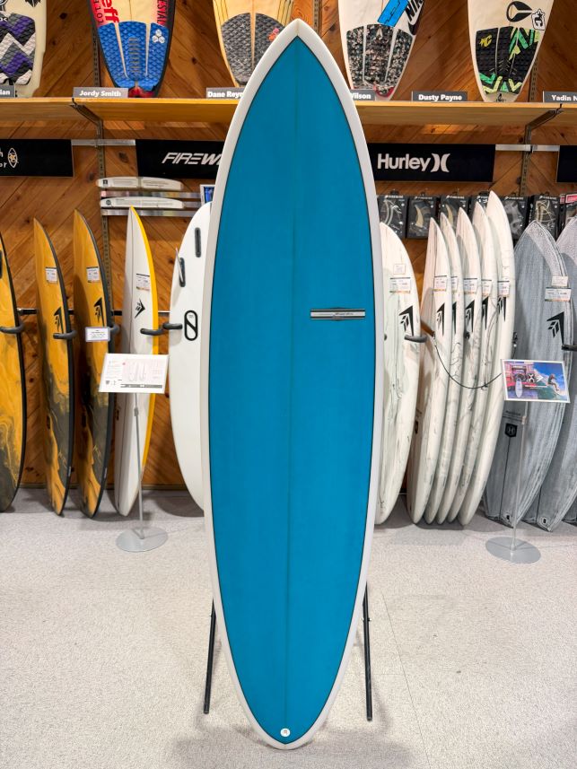IMG_9516 - SHOP NEWS | ムラサキスポーツオンラインストア 通販 Blue surfboard on a stand in a shop, surrounded by assorted surfboards on racks behind it.