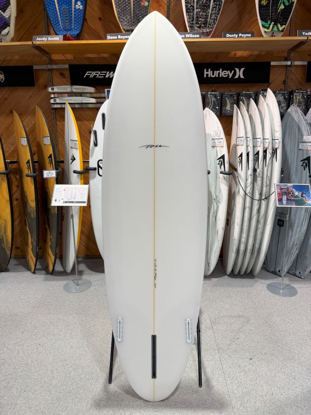 IMG_9518 - SHOP NEWS | ムラサキスポーツオンラインストア 通販 White surfboard on a display stand in a shop, with other boards on racks behind it.