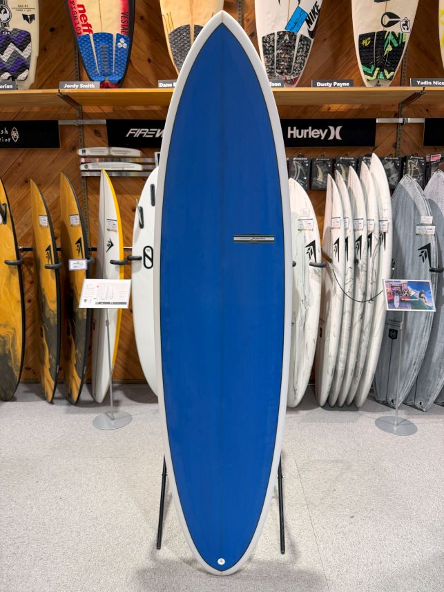 IMG_9521 - SHOP NEWS | ムラサキスポーツオンラインストア 通販 Blue surfboard on a stand in a surf shop display, with other boards lined up behind it on racks.