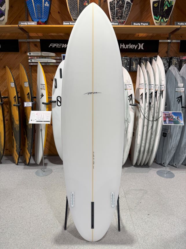 IMG_9522 - SHOP NEWS | ムラサキスポーツオンラインストア 通販 White shortboard on a display stand in a surf shop, surrounded by other boards on racks behind it.