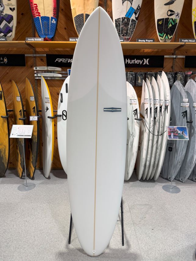 IMG_9525 - SHOP NEWS | ムラサキスポーツオンラインストア 通販 White shortboard surfboard on a display stand in a surf shop, with other boards visible in the background.