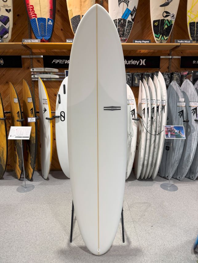 IMG_9529 - SHOP NEWS | ムラサキスポーツオンラインストア 通販 White surfboard on a stand in a surf shop, surrounded by other boards on racks and shelves.