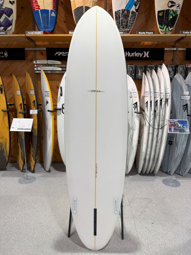 IMG_9530 - SHOP NEWS | ムラサキスポーツオンラインストア 通販 White surfboard with a thin gold stringer on a display stand in a surf shop, surrounded by other boards