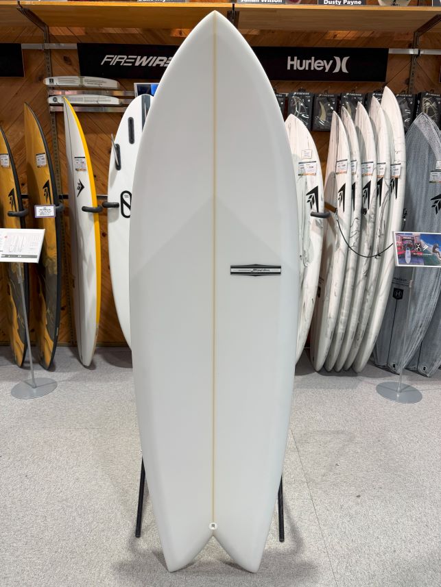 IMG_9533 - SHOP NEWS | ムラサキスポーツオンラインストア 通販 White surfboard displayed upright on a rack in a surf shop, surrounded by other boards and signage.