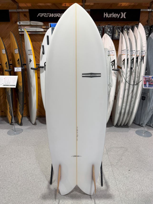 IMG_9534 - SHOP NEWS | ムラサキスポーツオンラインストア 通販 White surfboard on a display stand in a surf shop, with other boards lined up behind it on wooden shelving.