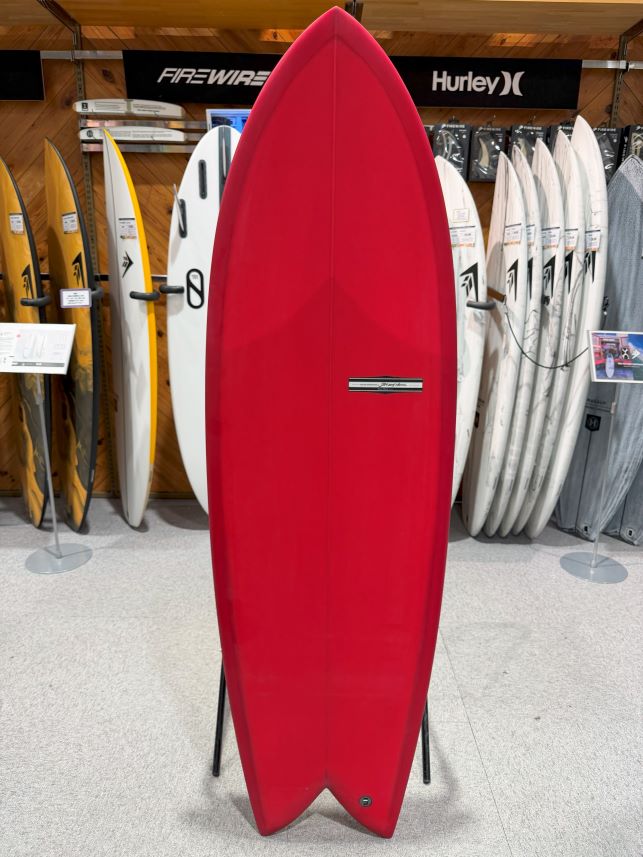 IMG_9537 - SHOP NEWS | ムラサキスポーツオンラインストア 通販 Red surfboard standing upright on a display rack in a surf shop, with other boards and brand banners visible in the background.