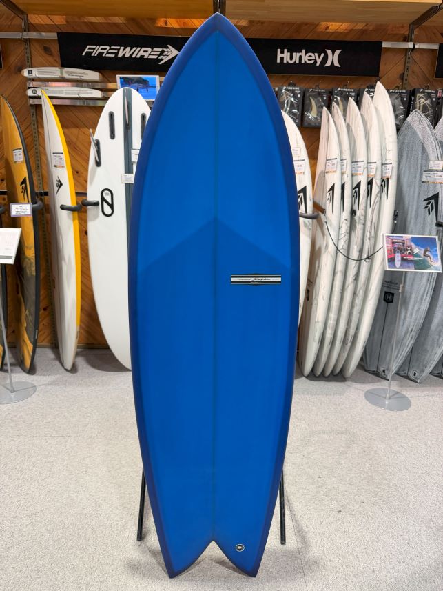 IMG_9541 - SHOP NEWS | ムラサキスポーツオンラインストア 通販 Blue shortboard surfboard on a stand in a store, surrounded by other boards and banners in the background.