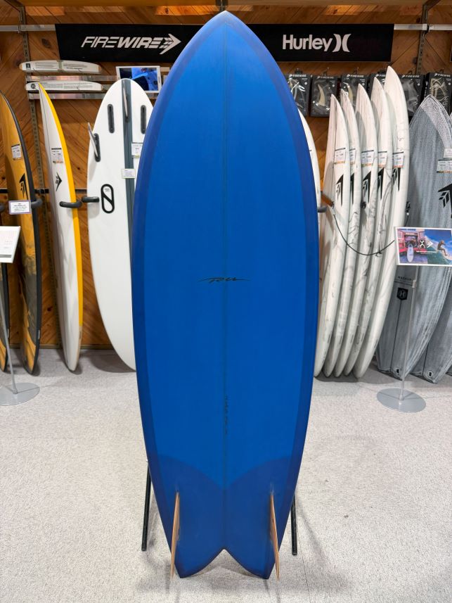 IMG_9542 - SHOP NEWS | ムラサキスポーツオンラインストア 通販 Blue surfboard on a stand in a surf shop, with white and patterned boards on racks in the background.