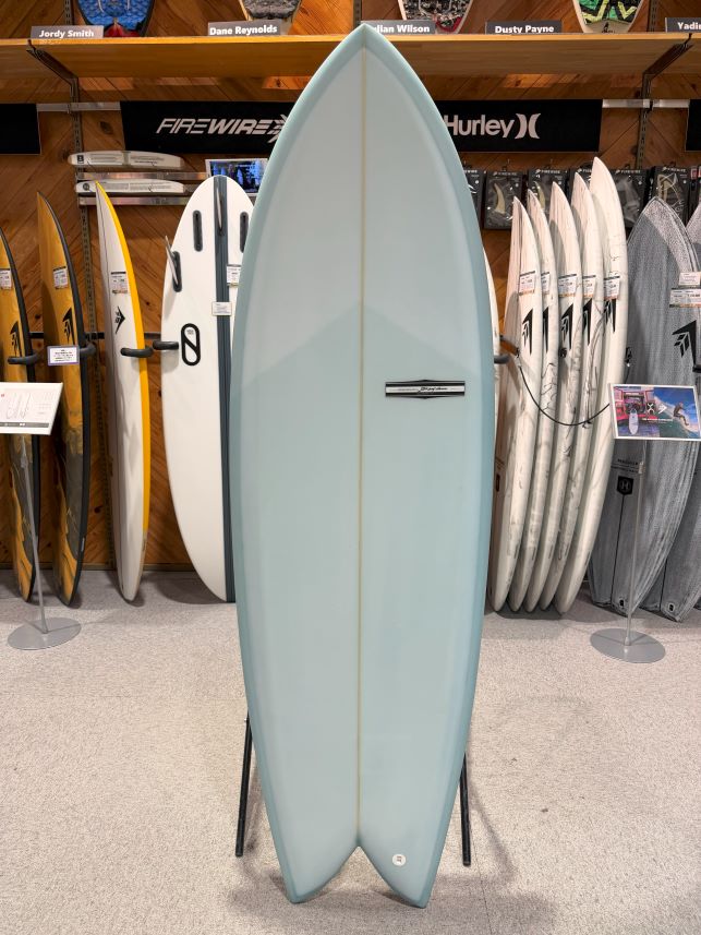 IMG_9545 - SHOP NEWS | ムラサキスポーツオンラインストア 通販 Light blue surfboard on a display stand in a surf shop, surrounded by other boards and shelves.