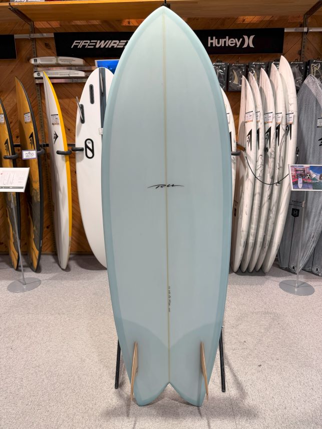 IMG_9546 - SHOP NEWS | ムラサキスポーツオンラインストア 通販 Light blue longboard surfboard on a display stand in a surf shop, with other boards and brands like Firewire and Hurley in the background.
