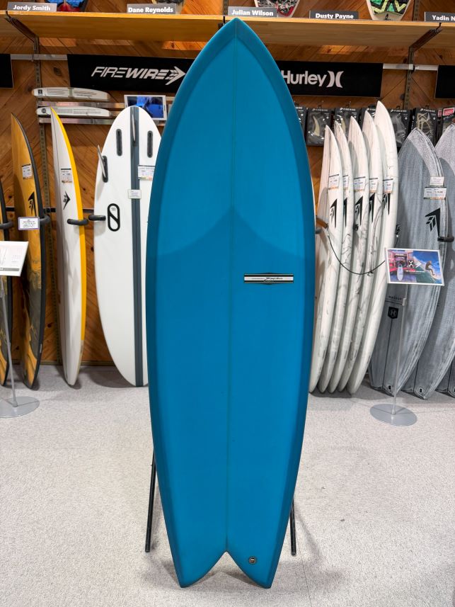 IMG_9548 - SHOP NEWS | ムラサキスポーツオンラインストア 通販 Tall blue surfboard on a display stand in a surf shop, with other boards lined up behind it.