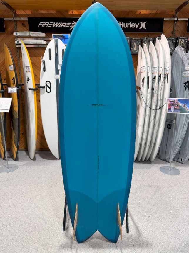 IMG_9549 - SHOP NEWS | ムラサキスポーツオンラインストア 通販 Blue surfboard standing upright on a display rack in a surf shop, with other boards and brand signage in the background.