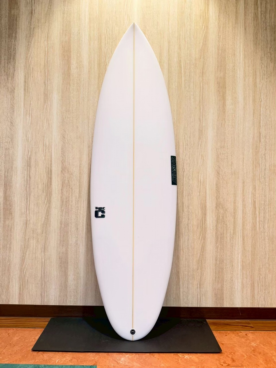LINE_ALBUM_2026 フリーダム_260601_3 - SHOP NEWS | ムラサキスポーツオンラインストア 通販 White surfboard standing upright against a wood-paneled wall on a black mat indoors, showing a central stringer and tail logo.
