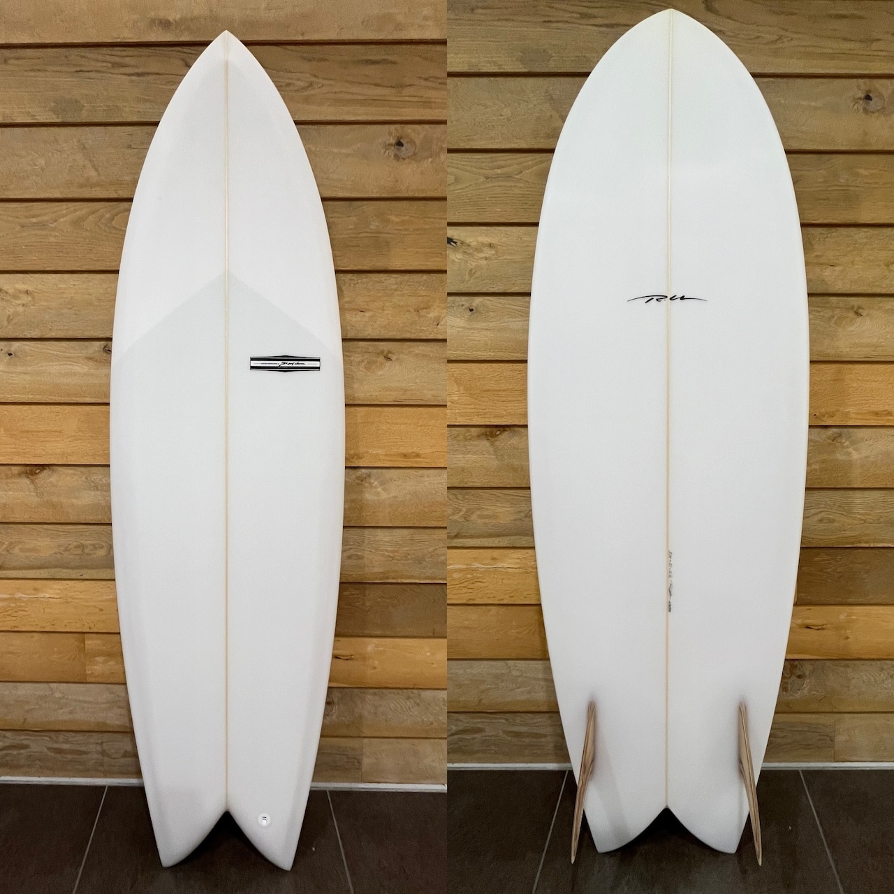 RU FISH CLR - SHOP NEWS | ムラサキスポーツオンラインストア 通販 Front and back views of a white shortboard propped against a wood wall; a small rectangular badge is on the front.