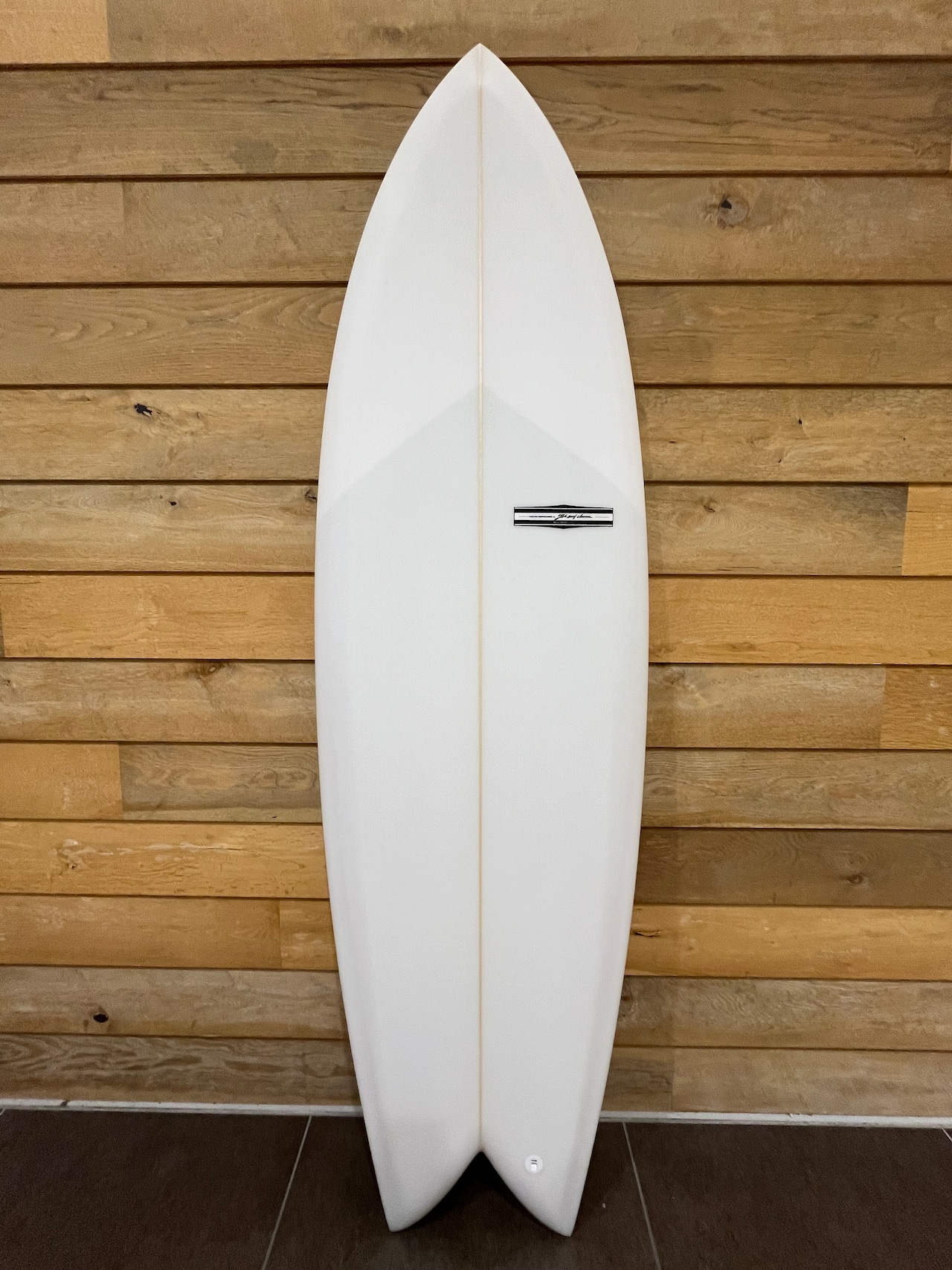 RU FISH1 - SHOP NEWS | ムラサキスポーツオンラインストア 通販 White surfboard standing upright against a wooden wall, with a central stringer and small logo plate.