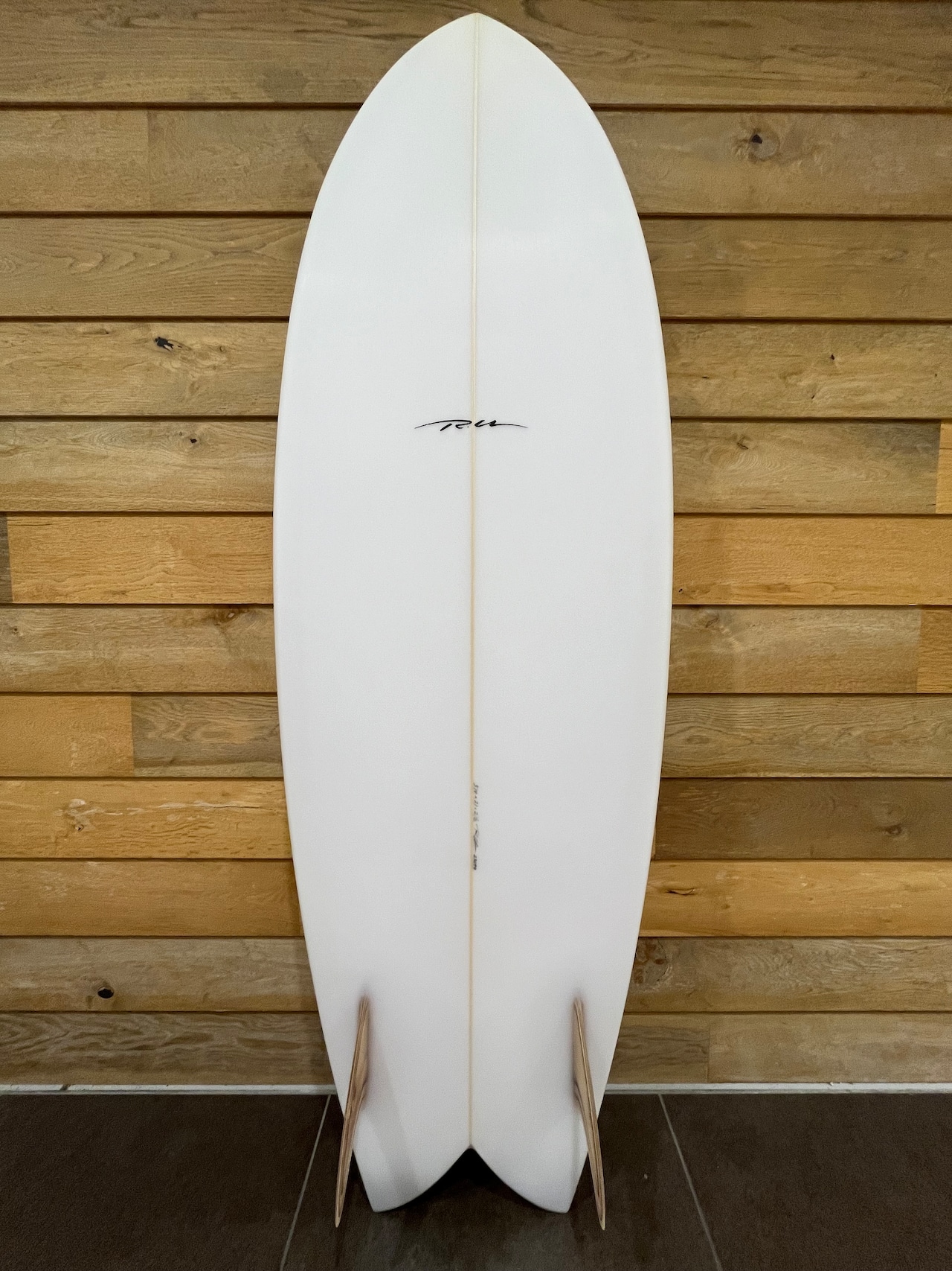 RU FISH2 - SHOP NEWS | ムラサキスポーツオンラインストア 通販 White surfboard standing upright against a wooden panel wall, with two light-colored fins at the tail end.