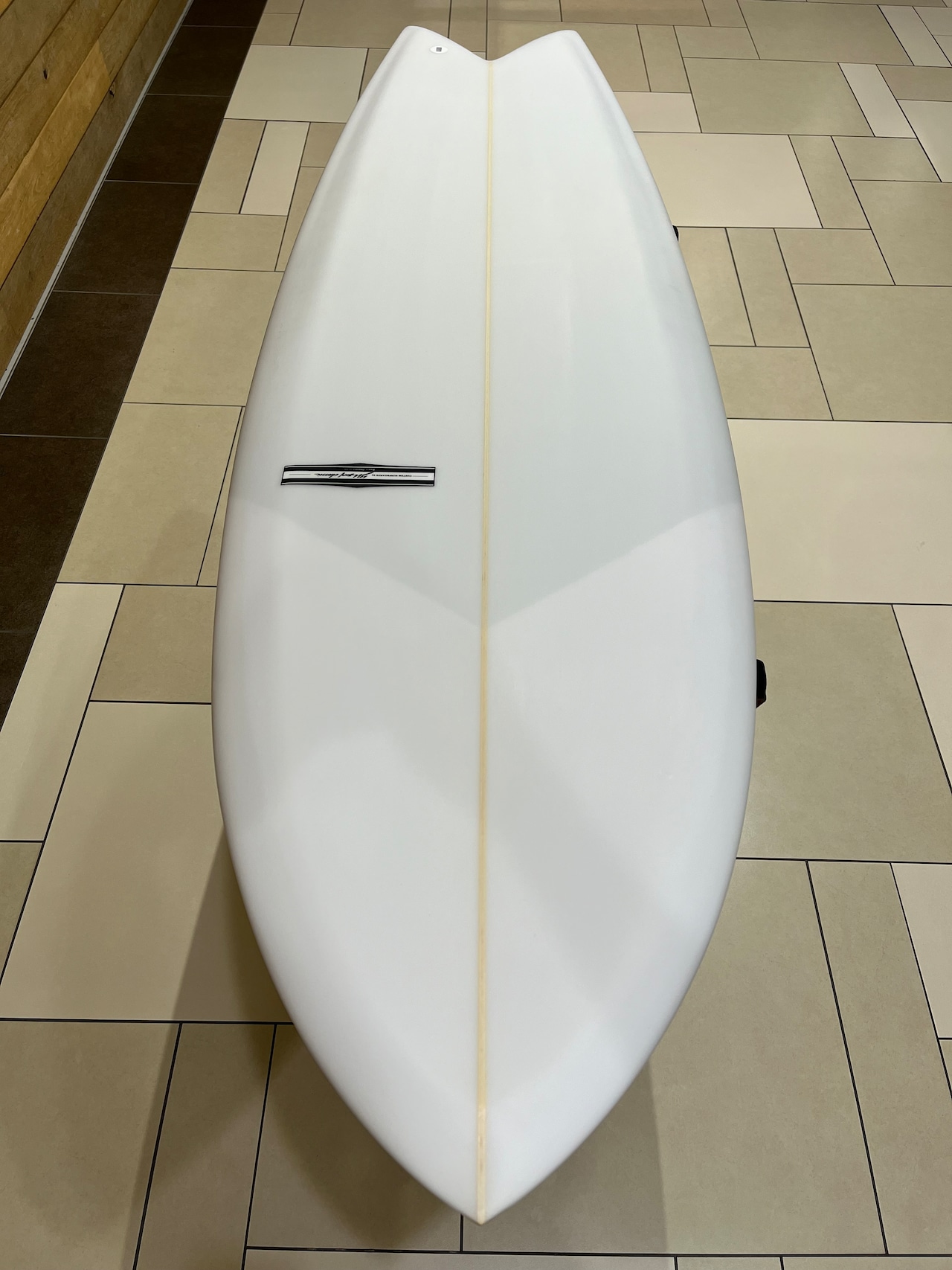 RU FISH3 - SHOP NEWS | ムラサキスポーツオンラインストア 通販 White surfboard viewed from above, lying on a beige tiled floor with a pale center stringer and a small black-and-white brand badge on the left side.
