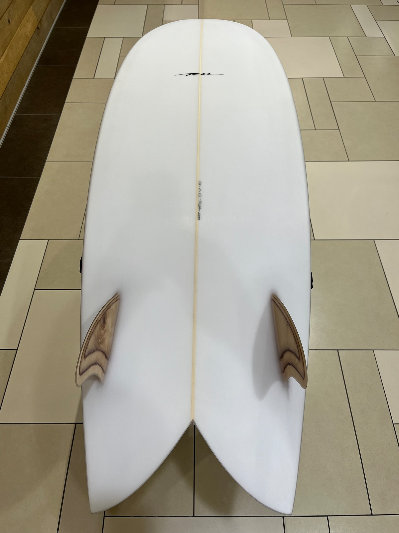 RU FISH4 - SHOP NEWS | ムラサキスポーツオンラインストア 通販 Top-down view of a white surfboard with a beige stringer and two wooden fins near the tail, resting on a tiled floor.