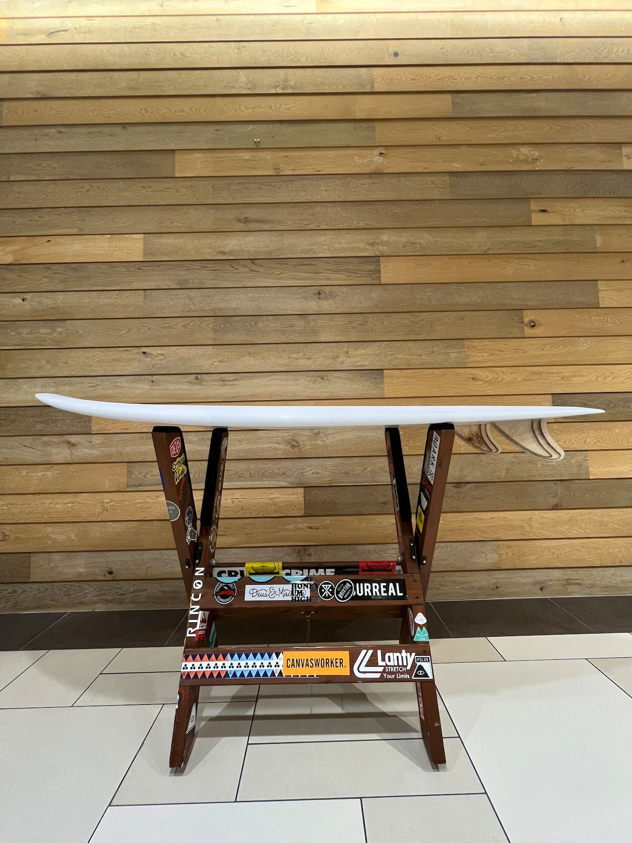 RU FISH5 - SHOP NEWS | ムラサキスポーツオンラインストア 通販 White surfboard resting on a sticker-covered wooden rack indoors against a light wood-paneled wall.