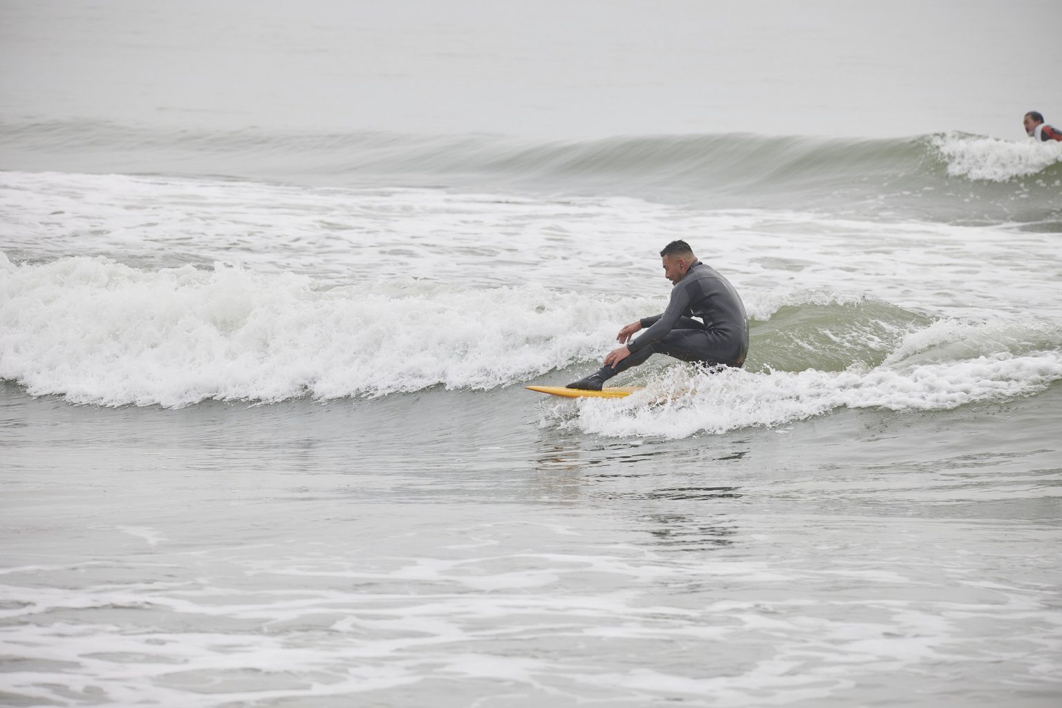 YU-SURF-CLASSIC-Arcana_1-1536x1024 - SHOP NEWS | ムラサキスポーツオンラインストア 通販 Surfer in a gray wetsuit riding a small wave on a yellow board; another surfer visible in the background.