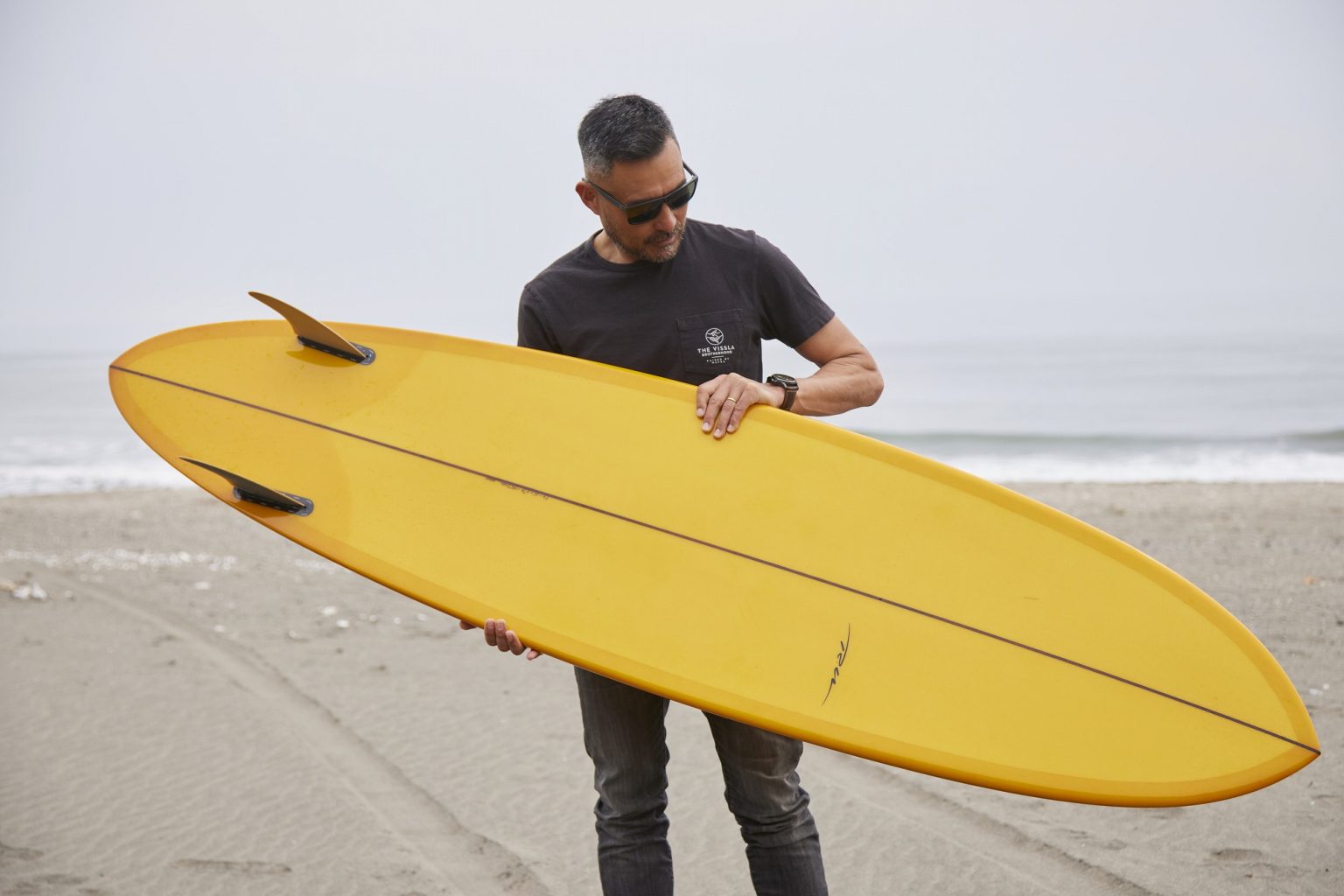YU-SURF-CLASSIC-Arcana_2-1536x1024 - SHOP NEWS | ムラサキスポーツオンラインストア 通販 Man on a beach holding a bright yellow surfboard across his chest, wearing sunglasses.