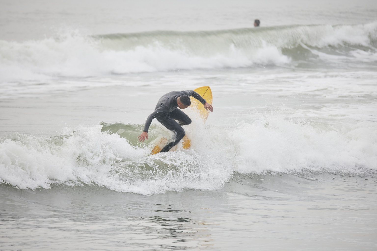 YU-SURF-CLASSIC-Arcana_5-1536x1024 - SHOP NEWS | ムラサキスポーツオンラインストア 通販 Surfer in a gray wetsuit rides a yellow surfboard on a small, frothy wave at the beach.