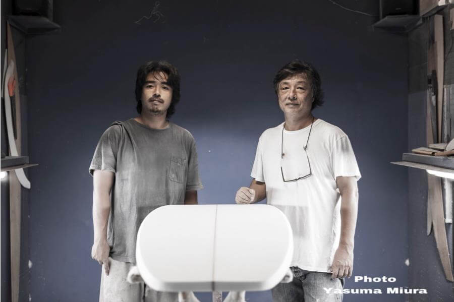 yu3 - SHOP NEWS | ムラサキスポーツオンラインストア 通販 Two men stand in a surfboard shaping shop beside a white surfboard blank, posing for the photo near a blue wall panelled workshop area with tools visible on the sides.
