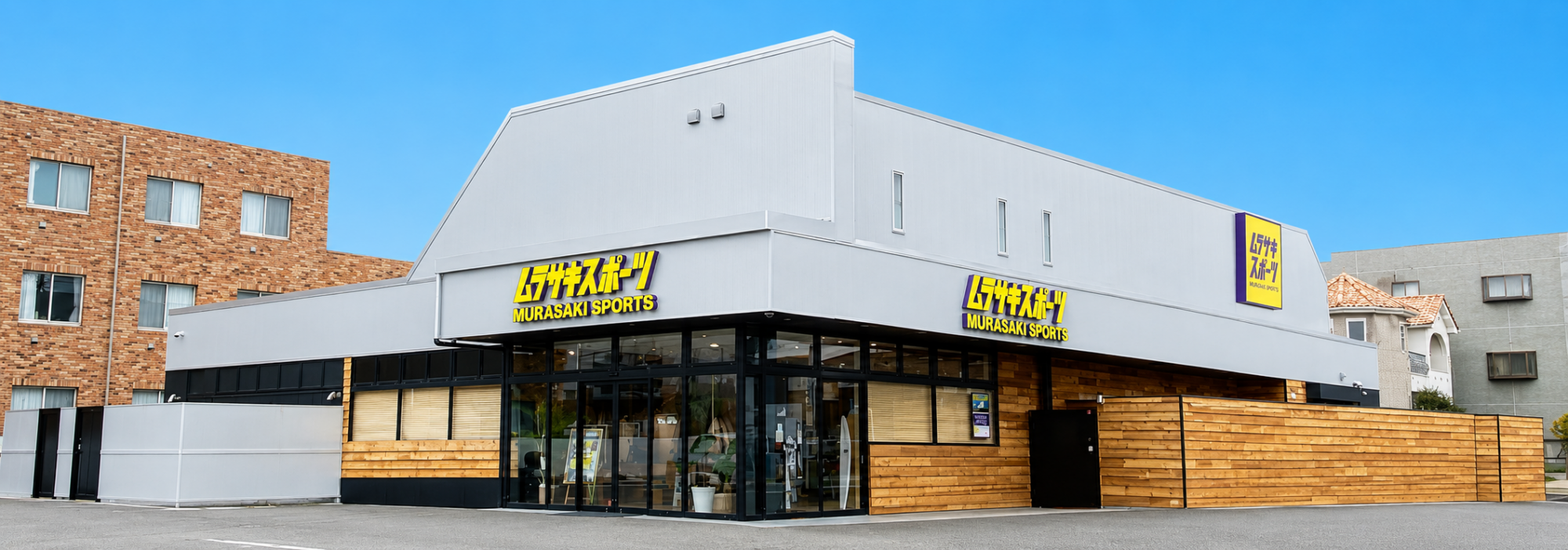 EE78B58C-12BD-477D-BDAA-89A85DB28EF2 - SHOP NEWS | ムラサキスポーツオンラインストア 通販 Murasaki Sports storefront with large glass windows, gray facade, and wooden fencing, under a clear blue sky.
