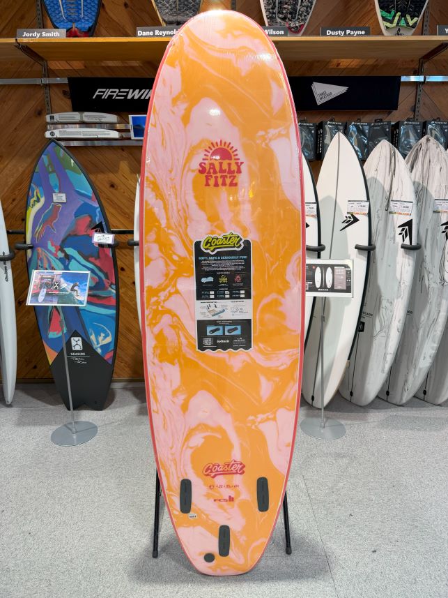 Orange-pink marbled surfboard on a stand in a shop, with Sally Fitz branding and a product info sticker.