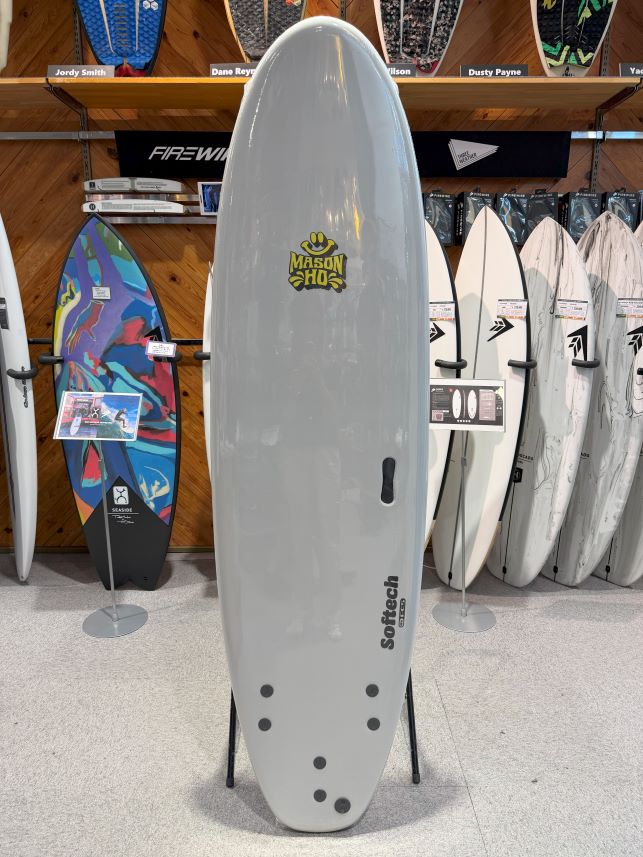 Gray surfboard on display in a shop, featuring a Mason Ho logo and Softech branding along the side. Other boards line the back wall.