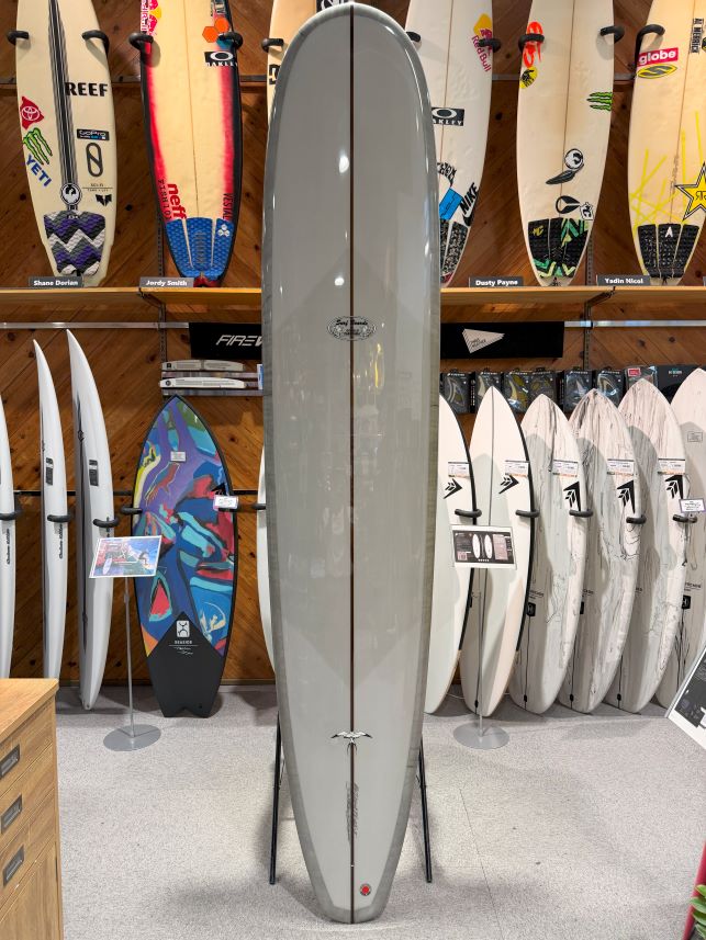 IMG_0264 - SHOP NEWS | ムラサキスポーツオンラインストア 通販 Tall gray surfboard on a stand displayed in a surf shop, with a row of boards and colorful stickers on the wall behind it.