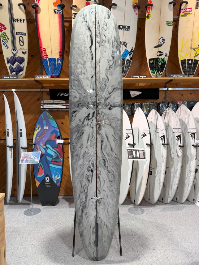 IMG_0266 - SHOP NEWS | ムラサキスポーツオンラインストア 通販 Marble-gray longboard on a stand in a surf shop, surrounded by other boards on display.