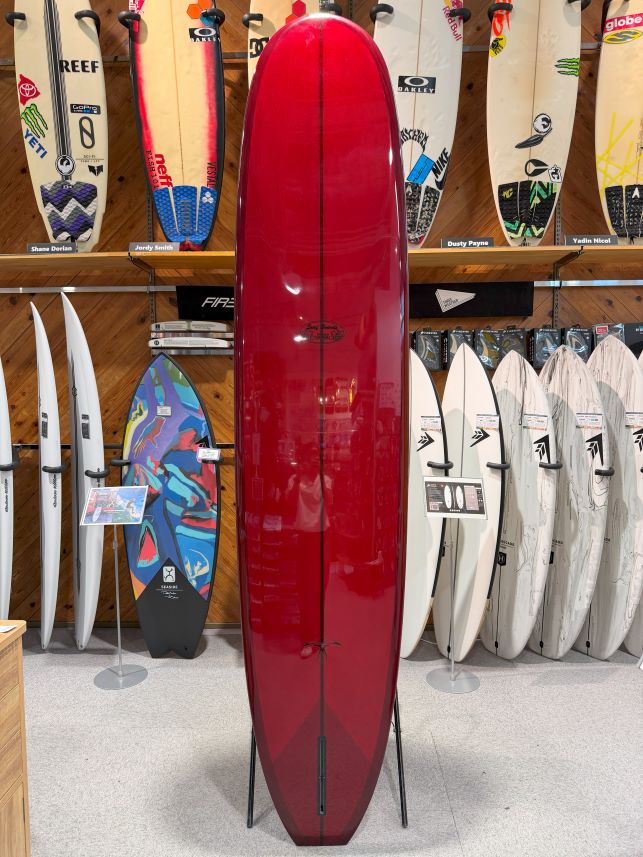 IMG_0270 - SHOP NEWS | ムラサキスポーツオンラインストア 通販 Red longboard surfboard on a stand in a surf shop, surrounded by various white and patterned boards on racks behind it.