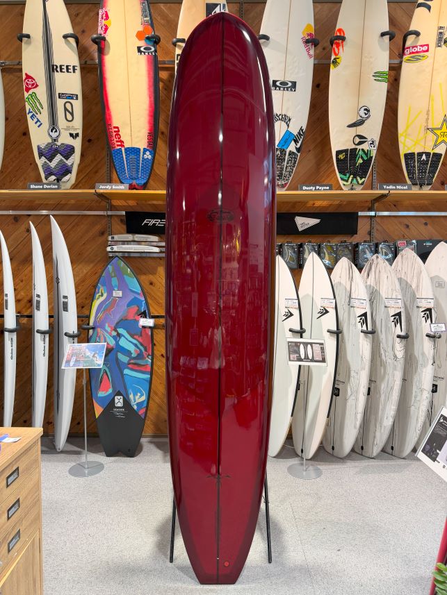 IMG_0274 - SHOP NEWS | ムラサキスポーツオンラインストア 通販 Red glossy surfboard on a stand in a shop display, with numerous boards lined along the walls behind.
