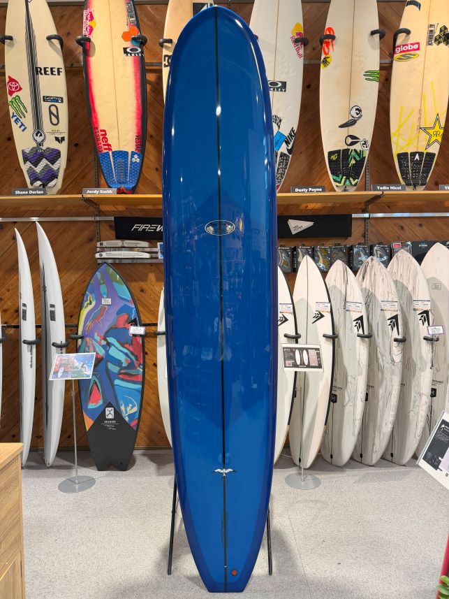 IMG_0278 - SHOP NEWS | ムラサキスポーツオンラインストア 通販 Blue longboard surfboard on a display rack in a surf shop, surrounded by other boards on shelves.